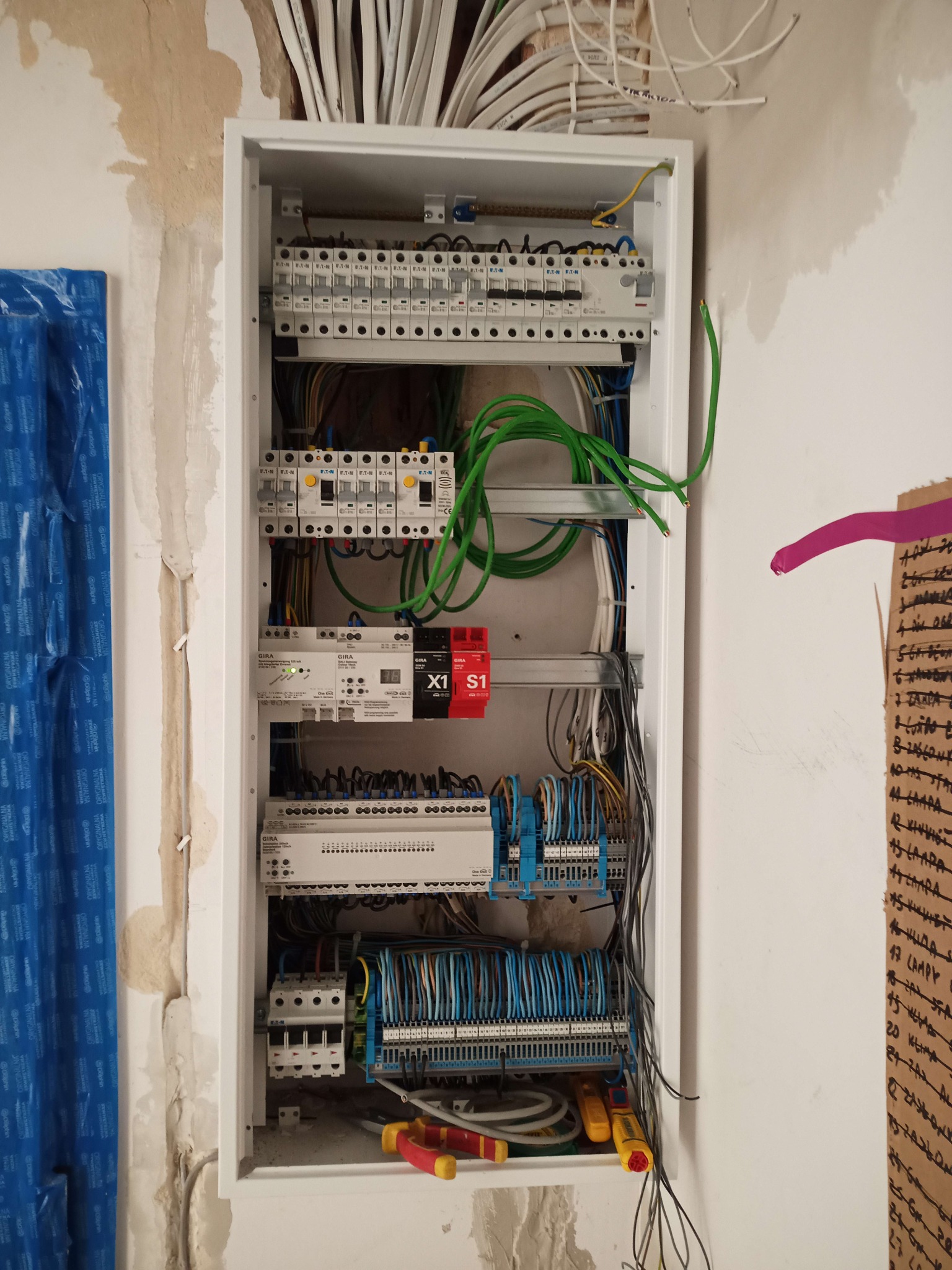 Rozdzielnica elektryczna w trakcie montażu, widoczne kable, bezpieczniki, moduły GIRA, narzędzia i prowizoryczne oznaczenia na ścianie.