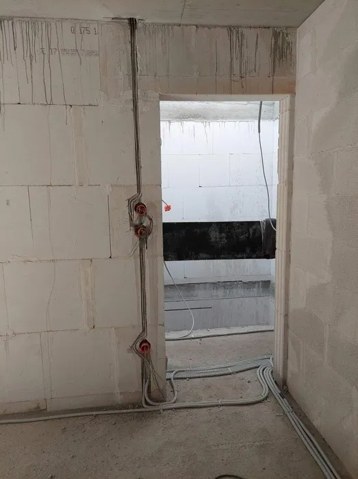 Instalacja elektryczna w surowym stanie, widoczne kable prowadzone po ścianie z bloczków, puszki instalacyjne z pomarańczowymi wnętrzami i peszle na podłodze.