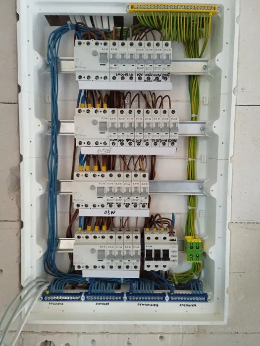 Rozdzielnica elektryczna z bezpiecznikami różnicowoprądowymi i listwami zaciskowymi, okablowanie w kolorach niebieskim, żółto-zielonym i brązowym, montaż w surowej ścianie z betonu komórkowego.