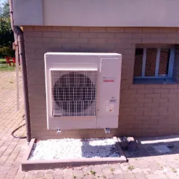 Zewnętrzna jednostka klimatyzacyjna Atlantic DC Inverter zamontowana na ścianie z cegły klinkierowej, z widocznym odpływem skroplin i ozdobnym podsypaniem z białego grysu.