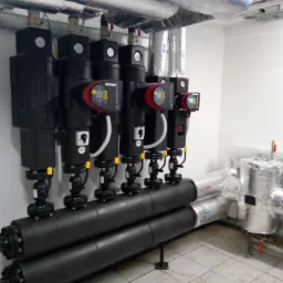 Rząd czarnych pomp obiegowych Grundfos z wyświetlaczami, podłączonych do zaizolowanych rur w pomieszczeniu technicznym z białą ścianą i szarą podłogą.