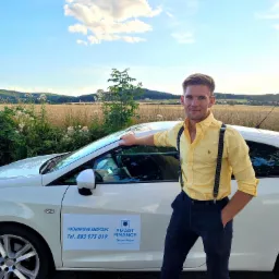 Uśmiechnięty mężczyzna w żółtej koszuli i szelkach opiera się o biały samochód z naklejką reklamową pośrednictwa kredytowego na tle krajobrazu z polami i wzgórzami.