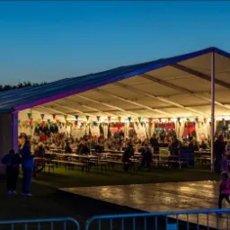 Duży, oświetlony namiot imprezowy z dekoracjami z chorągiewek, wypełniony gośćmi siedzącymi przy drewnianych stołach i ławkach, scena taneczna na pierwszym planie, wieczorne niebo w tle.