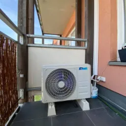 Biała jednostka zewnętrzna klimatyzacji zamontowana na balkonie, widoczne przewody i wiaderko farby, balkon wyłożony ciemnymi płytkami.