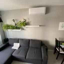 Biała jednostka klimatyzacyjna zamontowana na szarej ścianie nad sofą w salonie z roślinami na półkach i minimalistycznymi obrazkami.