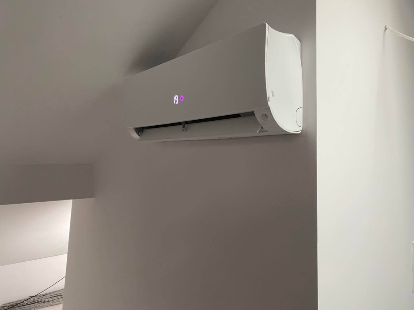Biała jednostka klimatyzacyjna zamontowana na szarej ścianie, wyświetlająca temperaturę 19 stopni Celsjusza, widok z dołu.
