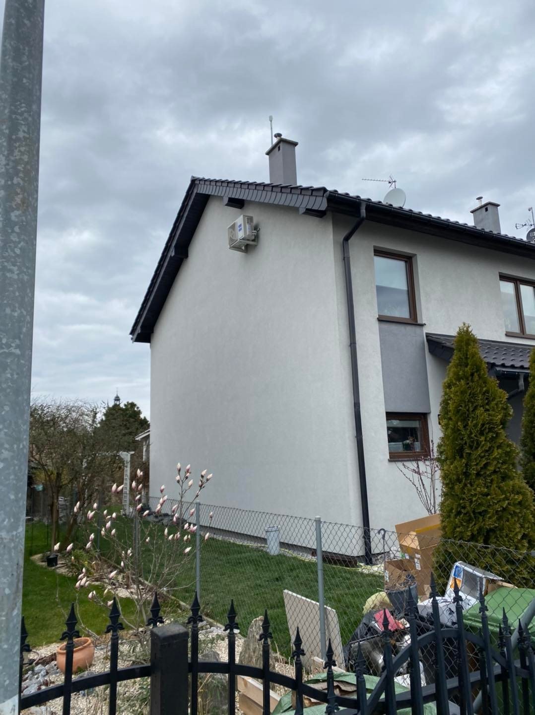 Boczny widok dwukondygnacyjnego domu z jasną elewacją, na której zamontowano jednostkę zewnętrzną systemu klimatyzacji; w tle pochmurne niebo i elementy ogrodu.