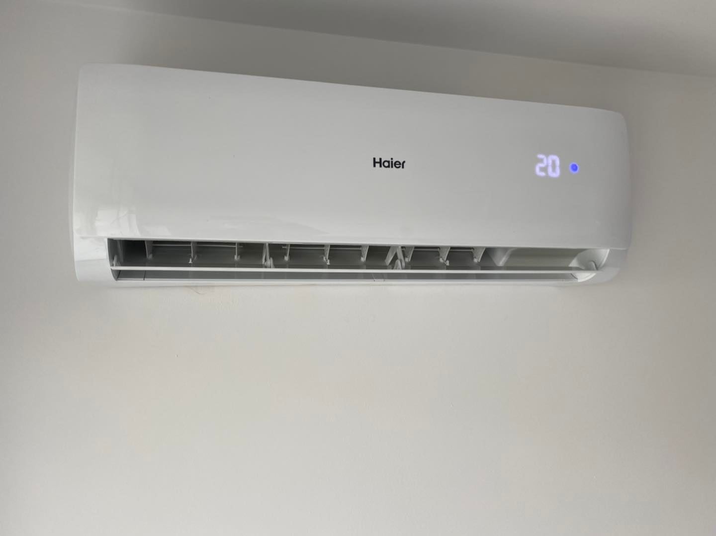 Biały klimatyzator Haier zamontowany na jasnej ścianie, wyświetlający temperaturę 20 stopni.
