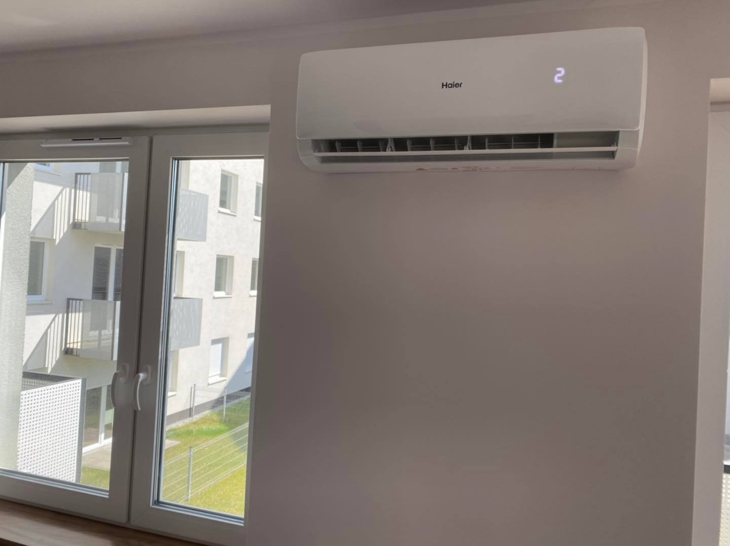 Klimatyzator Haier zamontowany na ścianie w mieszkaniu, widok z okna na budynek wielorodzinny z balkonami.