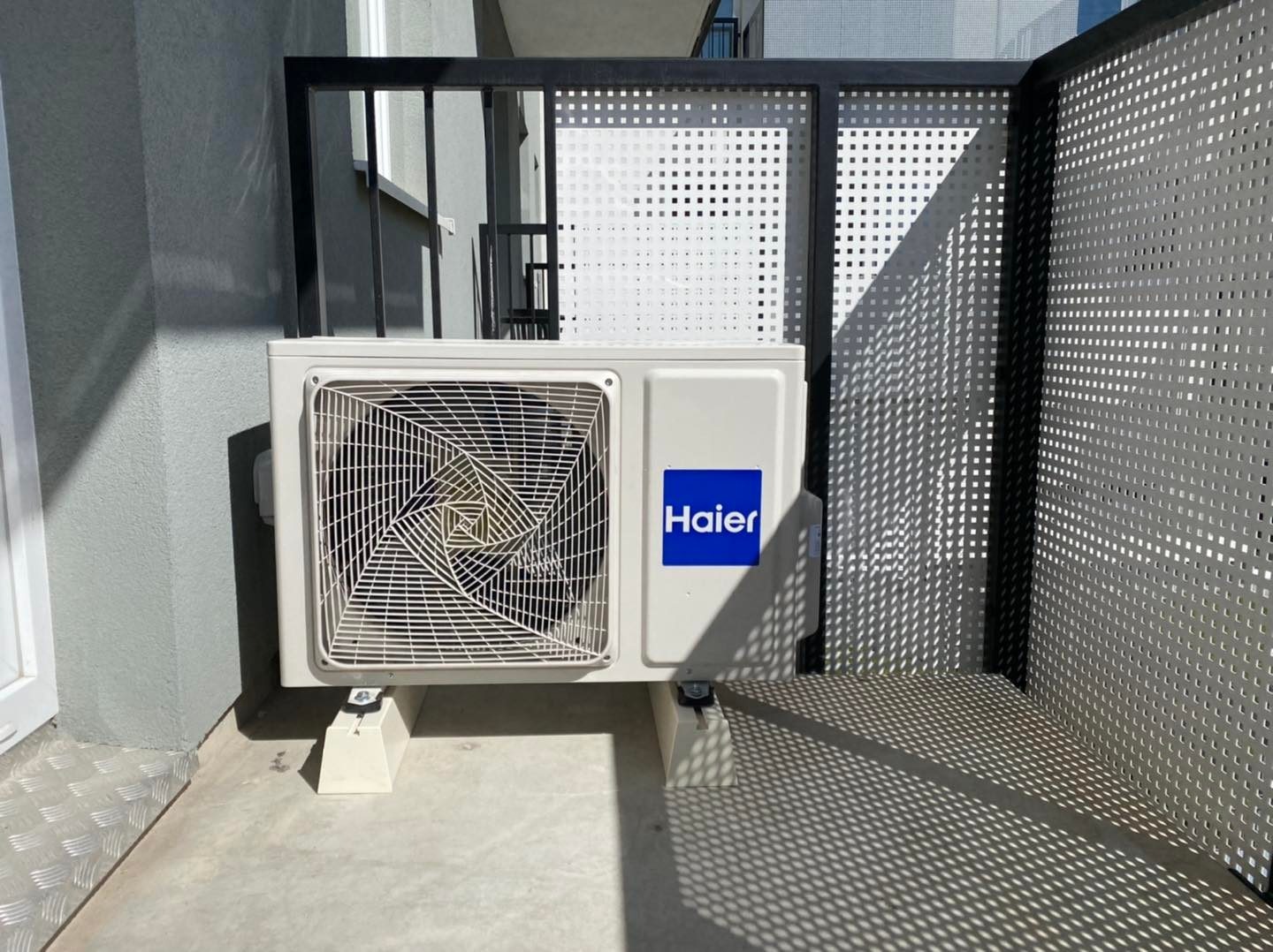 Biała jednostka zewnętrzna klimatyzatora Haier zamontowana na betonowym balkonie, z widoczną perforowaną barierką i cieniem rzucanym przez nią na podłogę.