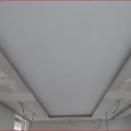 Niedokończony sufit podwieszany z widocznymi przewodami elektrycznymi i śladami po szpachlowaniu na obniżonych fragmentach, zarys okna w tle.