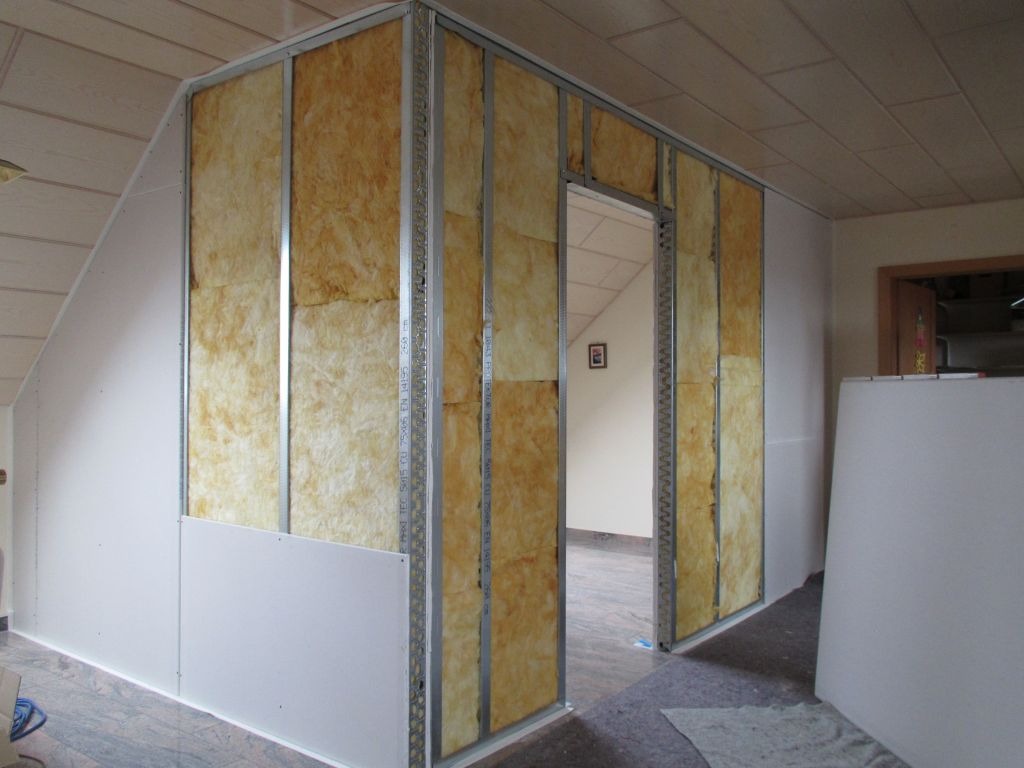 Wnętrze poddasza w trakcie adaptacji: widoczne metalowe profile, wełna mineralna w przestrzeni między nimi oraz częściowo zamontowane białe płyty. W tle widoczne kolejne pomieszczenie.