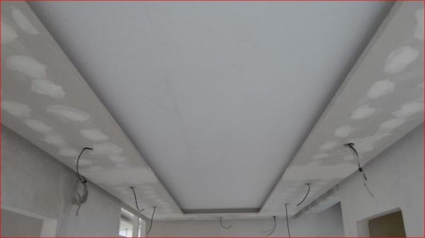 Niedokończony sufit podwieszany z widocznymi przewodami elektrycznymi i śladami po szpachlowaniu na obniżonych fragmentach, zarys okna w tle.