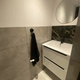 Nowoczesna łazienka z umywalką nablatową na szafce z dwiema szufladami, okrągłe lustro i szare płytki ścienne z czarnym marmurem za umywalką.