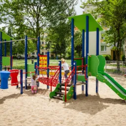 Kolorowy, nowoczesny zestaw zabaw dla dzieci z drabinkami, zjeżdżalnią i elementami wspinaczkowymi na piaszczystym podłożu; w tle budynek mieszkalny i kobieta siedząca na ławce.