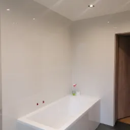 Jasna łazienka z wanną, wykończona białymi płytkami, z punktowym oświetleniem sufitowym i drewnianymi drzwiami po prawej stronie.