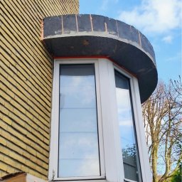 B&B Gw Sp Zoo - Wystający wykusz okienny z oknem PCV podczas budowy, z widocznymi elementami murarskimi i żółtą cegłą klinkierową na elewacji budynku. Na pierwszym planie kartonowe pudło.