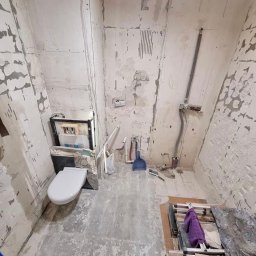 B&B Gw Sp Zoo - Łazienka w trakcie remontu: widoczne niedokończone ściany, zamontowany stelaż podtynkowy WC z częściowo założoną miską, narzędzia oraz pozostałości po skuciu płytek na podłodze.