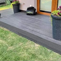 B&B Gw Sp Zoo - Szary taras kompozytowy z donicami kwiatów, przylegający do domu z drewnianymi drzwiami balkonowymi i zielonym trawnikiem.