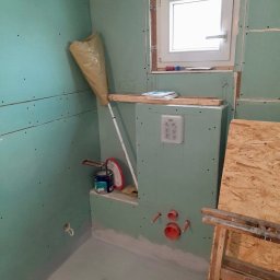 B&B Gw Sp Zoo - Łazienka w trakcie remontu: ściany pokryte płytami gipsowo-kartonowymi, widoczne instalacje wodne i kanalizacyjne, puszka podtynkową WC, okno, pędzel, wiadro z farbą i drabina.