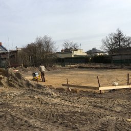 B&B Gw Sp Zoo - Przygotowanie terenu pod budowę: pracownik zagęszcza ziemię za pomocą zagęszczarki wibracyjnej, widoczne wytyczone granice przyszłego budynku za pomocą drewnianych kołków i listew, w tle zabudowa...
