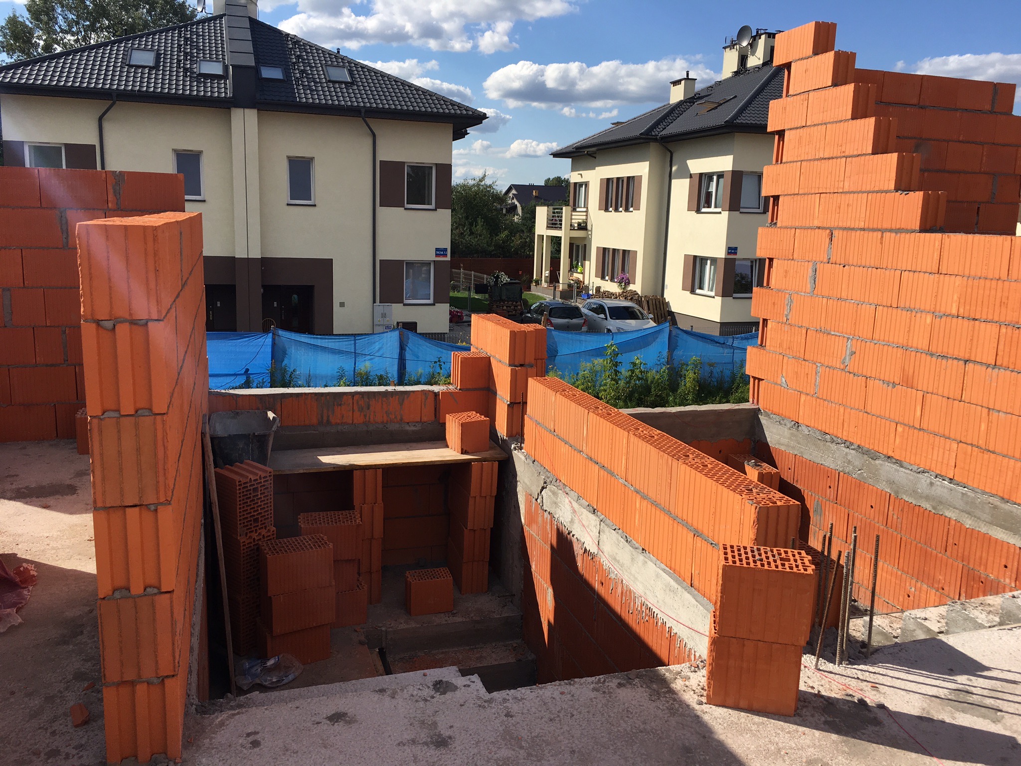 Wznoszenie ścian z czerwonej ceramiki porotherm na budowie domu, widoczne zbrojenia i sąsiednie budynki w tle.