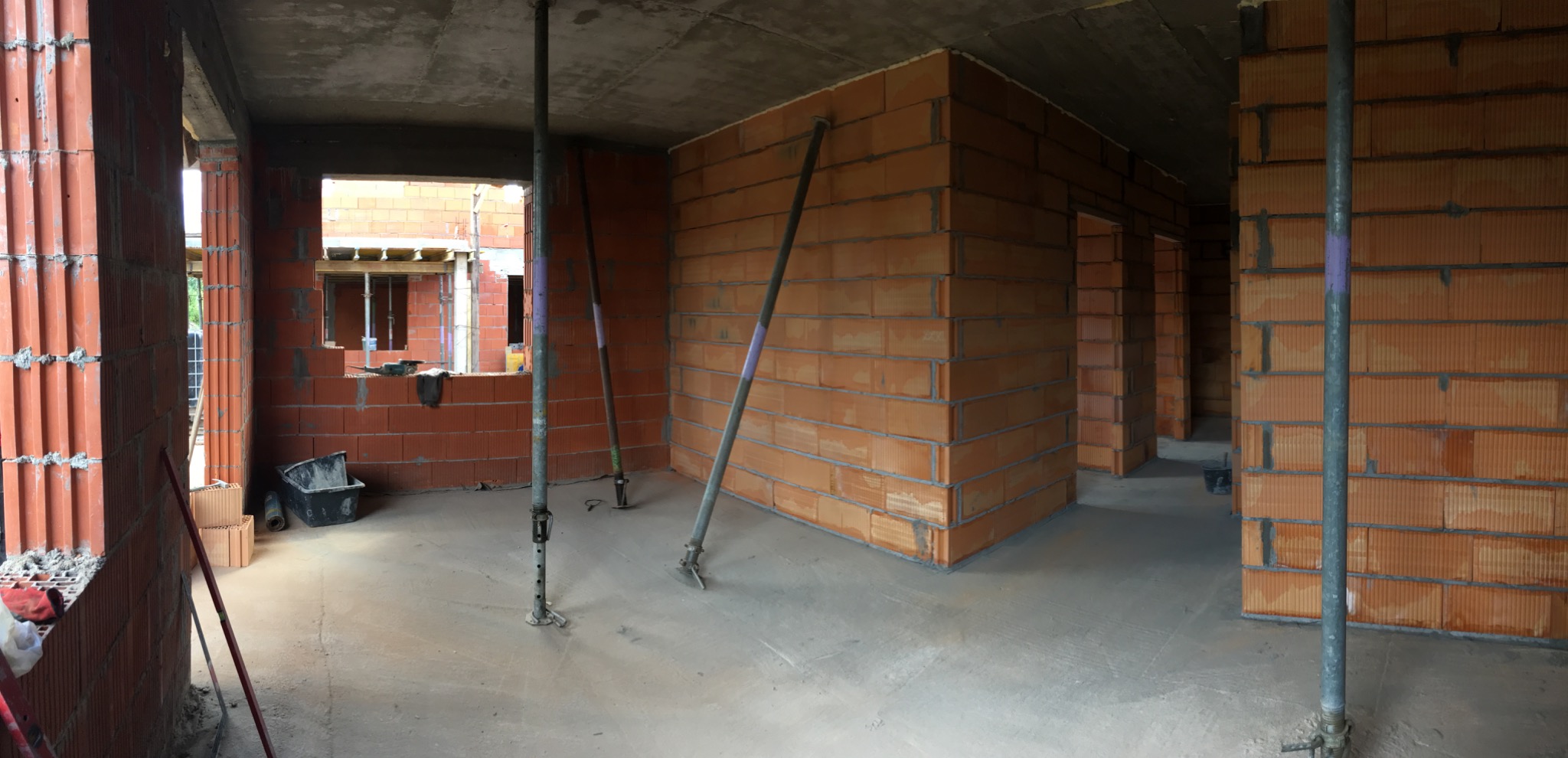 Wnętrze domu murowanego w trakcie budowy: ściany z czerwonej cegły ceramicznej, metalowe podpory stropowe, surowy betonowy strop, widok na kolejne pomieszczenia.