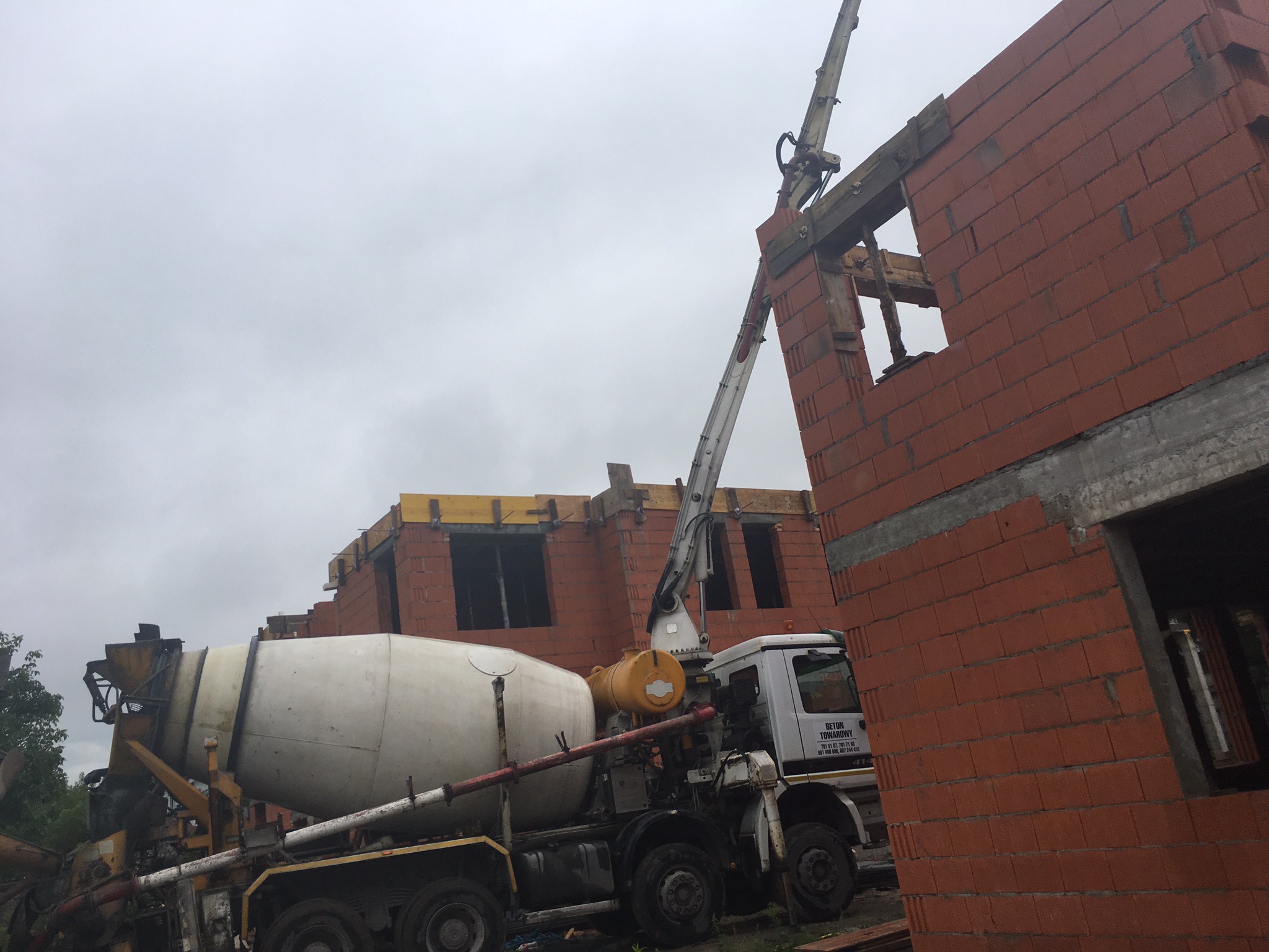 Betoniarka z wysięgnikiem podaje beton na wznoszone ściany z czerwonej cegły dziurawki na budowie domu, pochmurne niebo w tle.