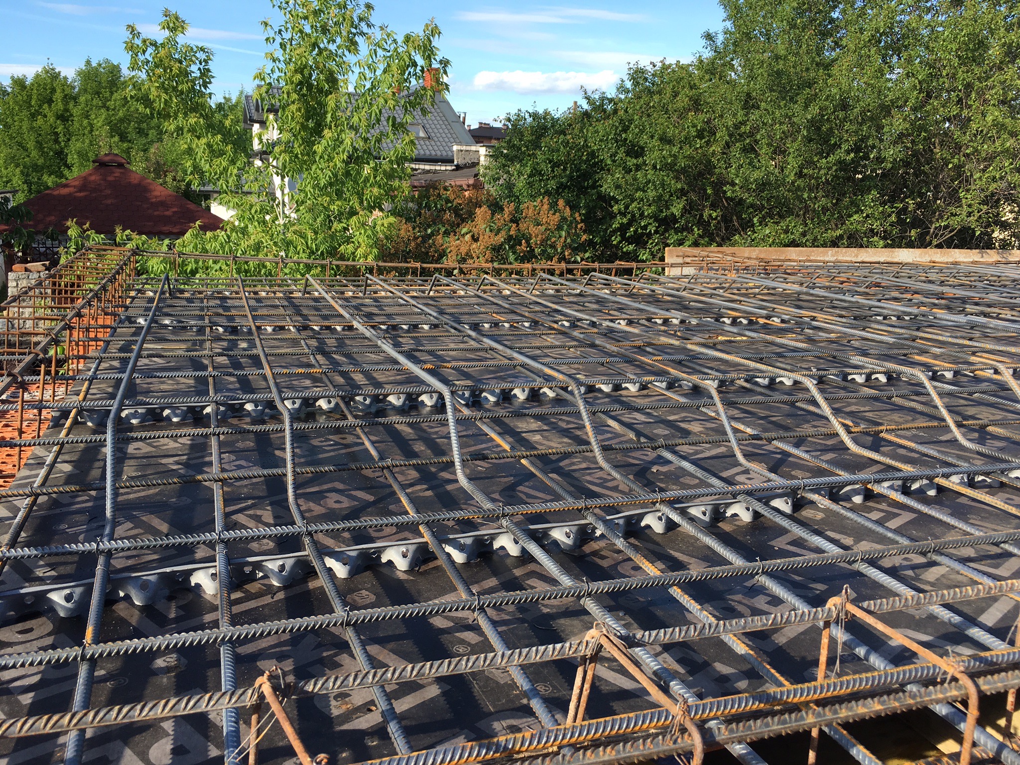 Zbrojenie płyty fundamentowej z dystansami z tworzywa sztucznego, widok z góry na tle zieleni i zabudowy jednorodzinnej.