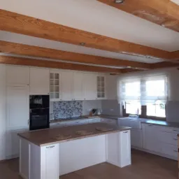 Jasna kuchnia z wyspą w Chyżnem, drewniane belki stropowe i białe fronty szafek.