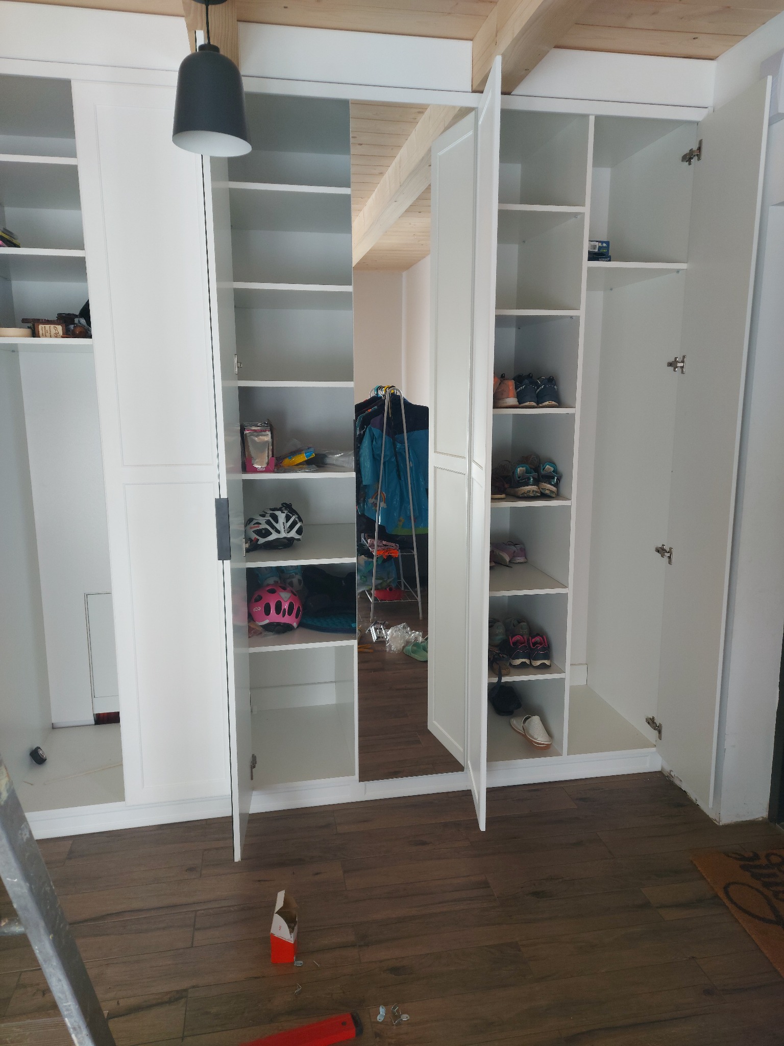Biała szafa wnękowa z otwartymi drzwiami, widoczne półki z butami i kaskami, drewniana podłoga i sufit, drabina oparta o bok szafy.