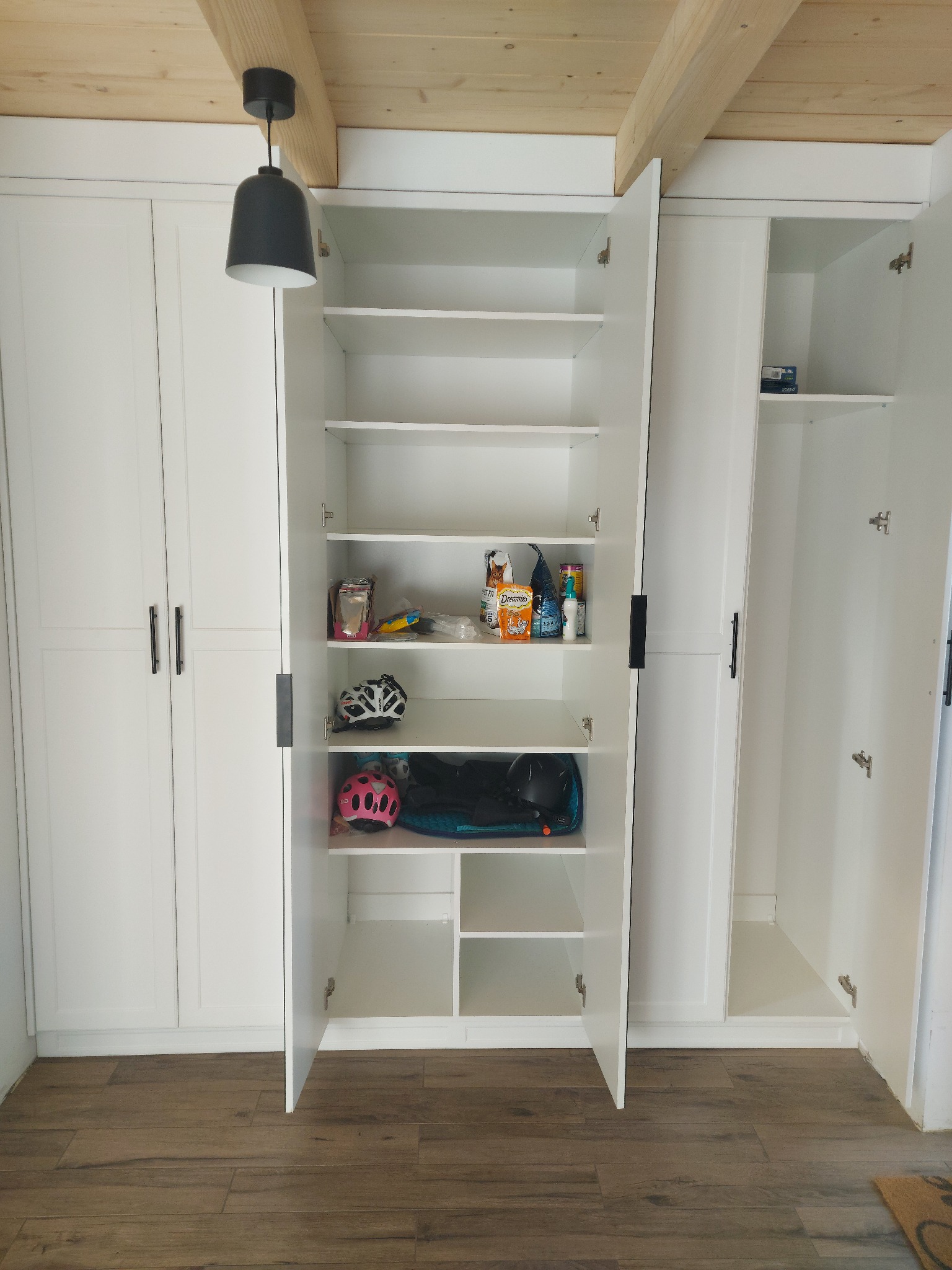 Biała szafa wnękowa z otwartymi drzwiami, widoczne półki z różnymi przedmiotami, w tym kaski rowerowe, na podłodze panele imitujące drewno, sufit z drewnianymi belkami.
