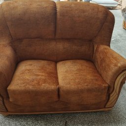 Zakład Usługowy Tapicerstwo Meblowe Jacek Sęków - Dwuosobowa sofa w odcieniach brązu z drewnianymi zdobieniami na podłokietnikach, widoczna w warsztacie tapicerskim z elementami pracy w tle.