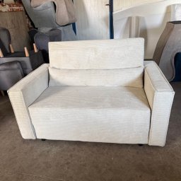 Zakład Usługowy Tapicerstwo Meblowe Jacek Sęków - Kremowy, dwuosobowy fotel tapicerowany sztruksem, z niskimi nóżkami, na tle warsztatu tapicerskiego z elementami innych mebli w trakcie renowacji.