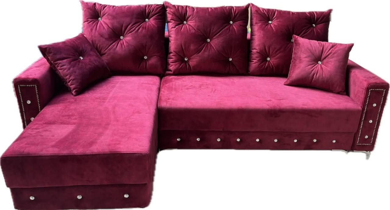 Bordowa sofa narożna z weluru, zdobiona kryształkami na poduszkach i bokach, na niskich nóżkach. Widok z przodu, na białym tle.