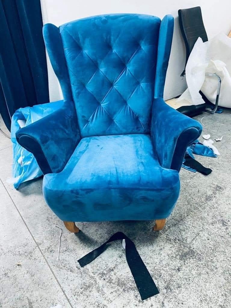 Błękitny fotel typu uszak z pikowanym oparciem, stojący na drewnianych nóżkach. Na podłodze leży czarny pasek materiału. W tle widoczne materiały i elementy meblowe.