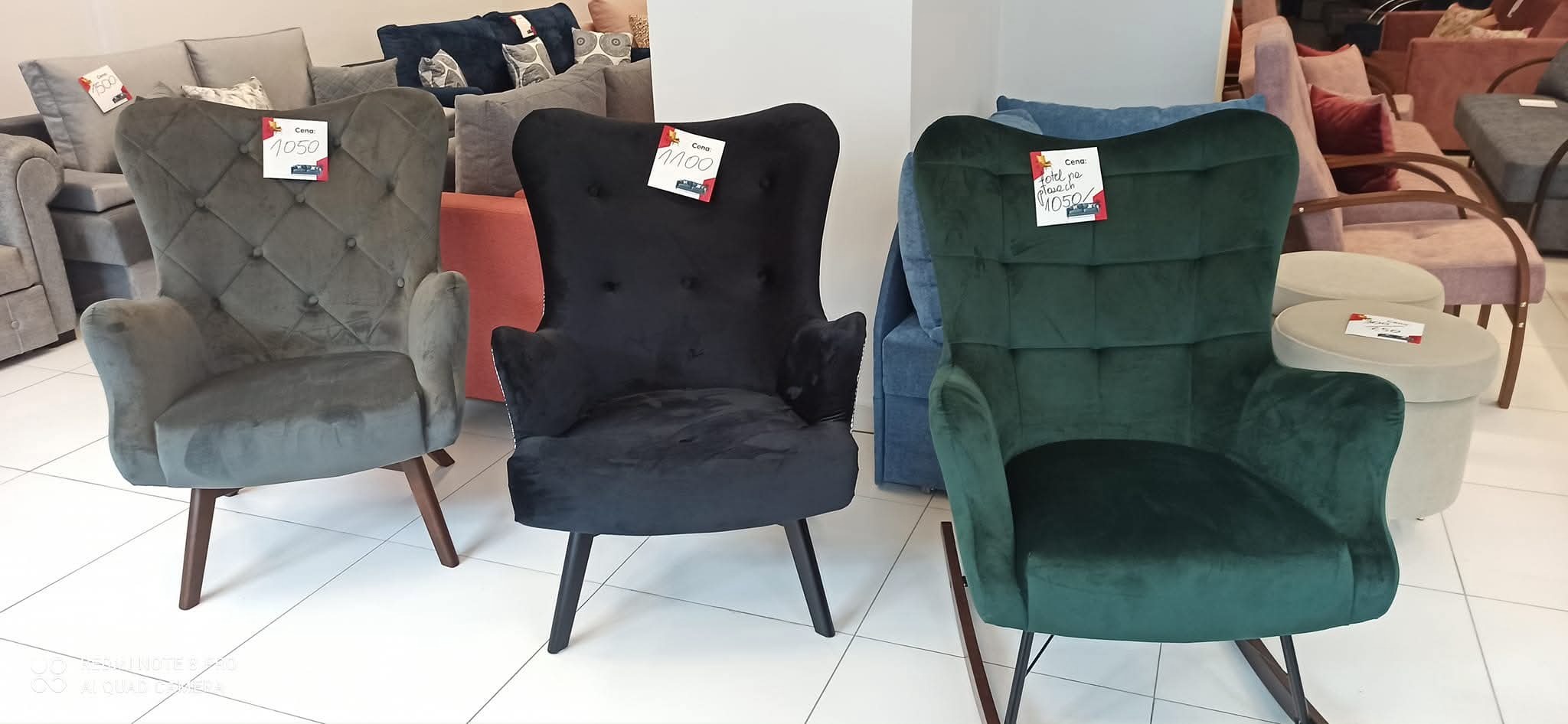 Trzy tapicerowane fotele w różnych kolorach (szary, czarny, zielony) z cenami, ustawione w salonie meblowym. Widoczne inne meble w tle.