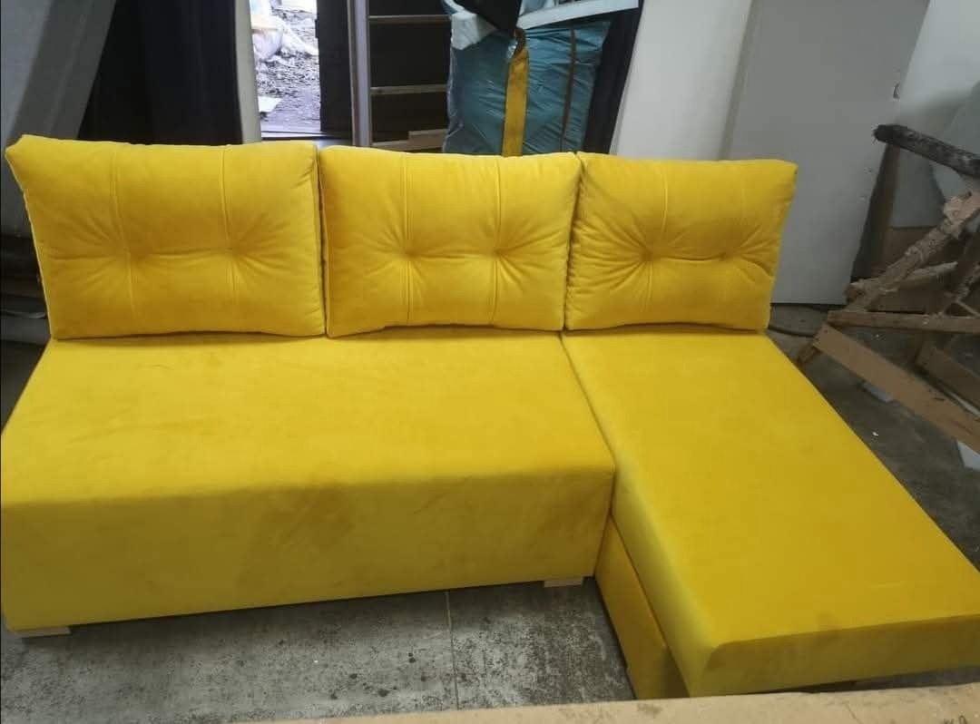 Żółta, narożna sofa z trzema poduszkami, wykonana z miękkiego weluru. Widoczne detale pikowania na poduszkach i minimalistyczne nóżki. Ujęcie z bliska, w warsztacie.