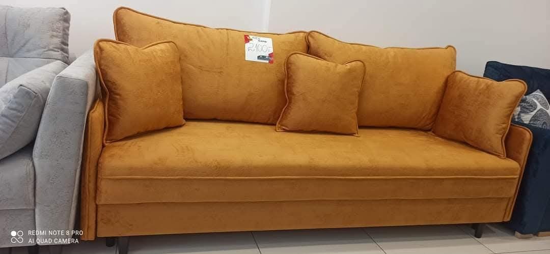 Musztardowa sofa z poduszkami, widoczna cena na jednej z nich. Obok fragment szarej sofy. Minimalistyczny design, gładka tkanina. Ujęcie z przodu, w neutralnym oświetleniu.
