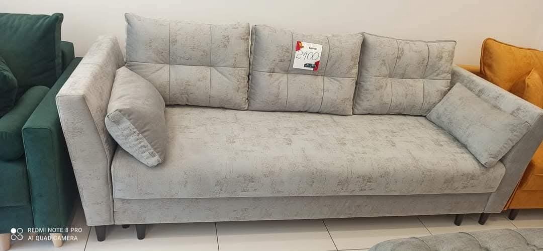 Jasnoszara sofa z poduszkami, z ceną 2100, na tle innych mebli w salonie. Widoczne detale tapicerki i nóżki sofy.
