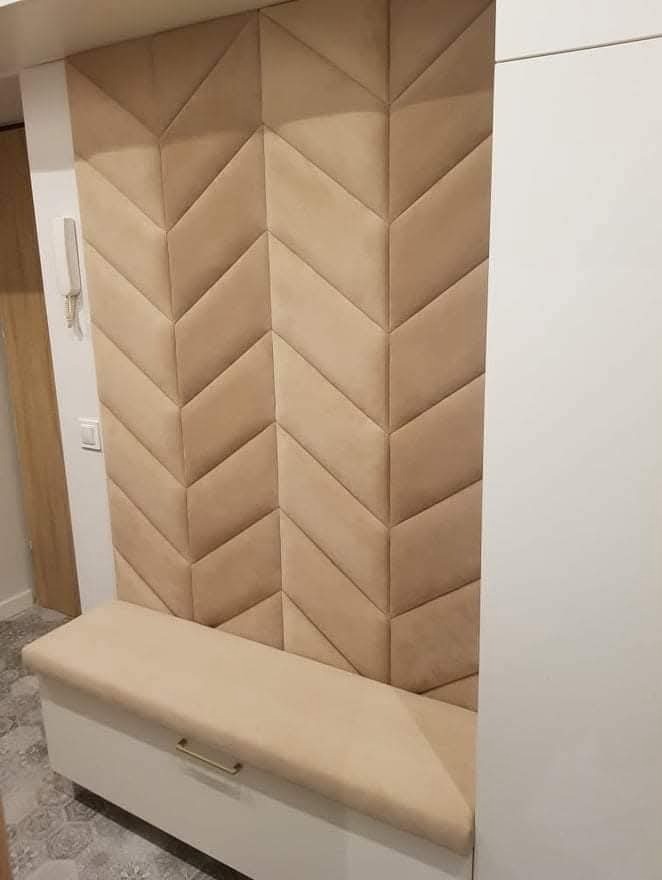 Beżowy panel tapicerowany w jodełkę, zintegrowany z siedziskiem i szafką w przedpokoju. Nowoczesny design, miękkie wykończenie, subtelne oświetlenie.