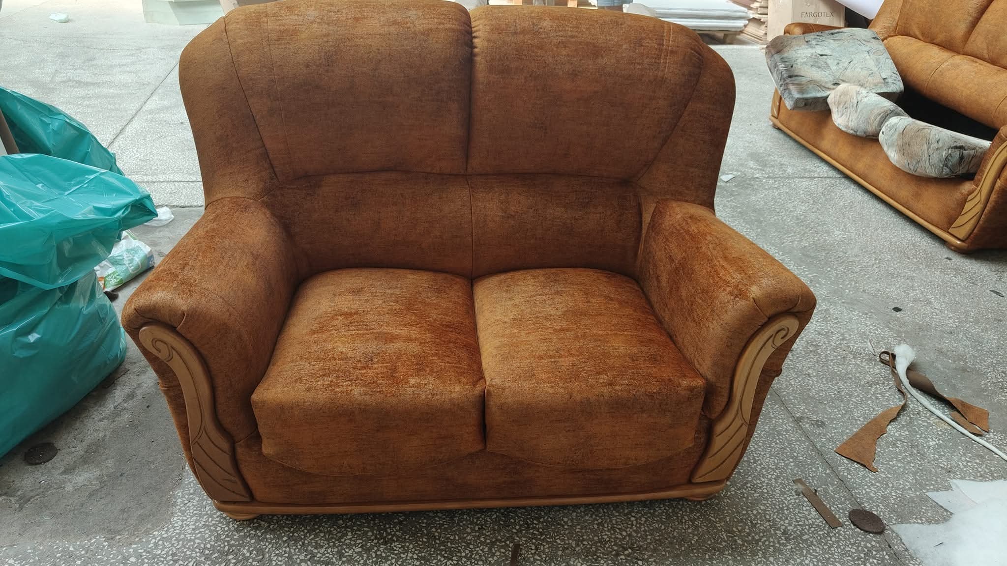 Dwuosobowa sofa w odcieniach brązu z drewnianymi zdobieniami na podłokietnikach, widoczna w warsztacie tapicerskim z elementami pracy w tle.