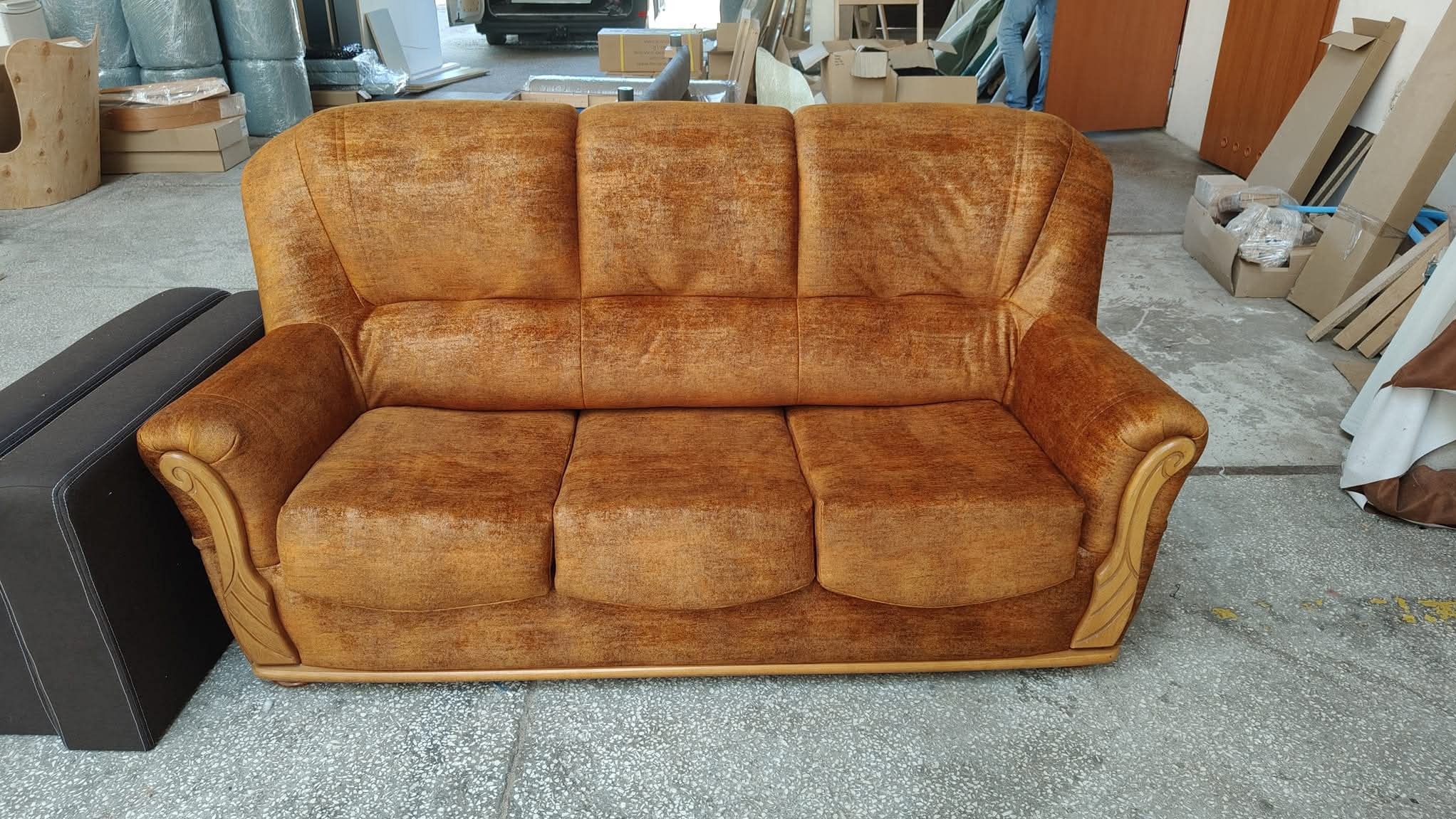 Pomarańczowa sofa z welurową tapicerką i drewnianymi zdobieniami na podłokietnikach, ustawiona w warsztacie tapicerskim, obok widoczny fragment innego mebla.