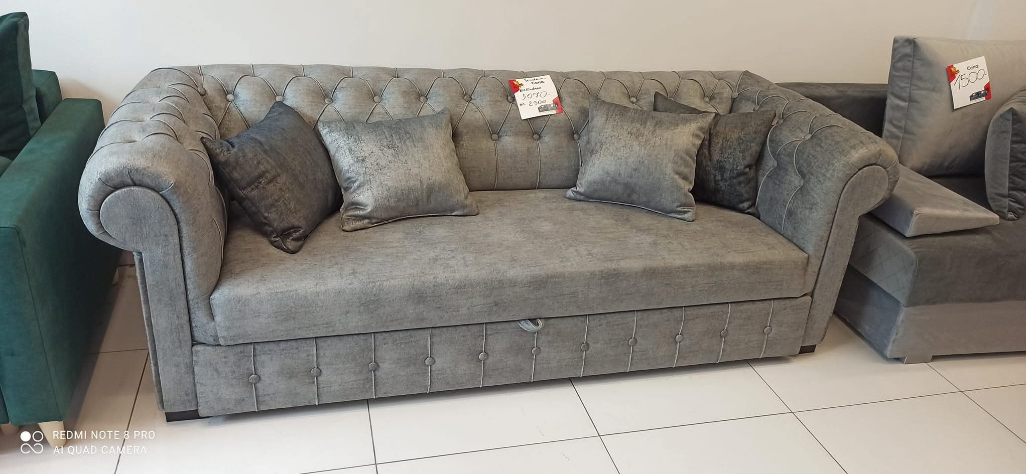 Szara sofa z poduszkami w stylu chesterfield, z widocznym mechanizmem rozkładania i cenówką. Obok fragment zielonej sofy. Ujęcie z bliska, w salonie.