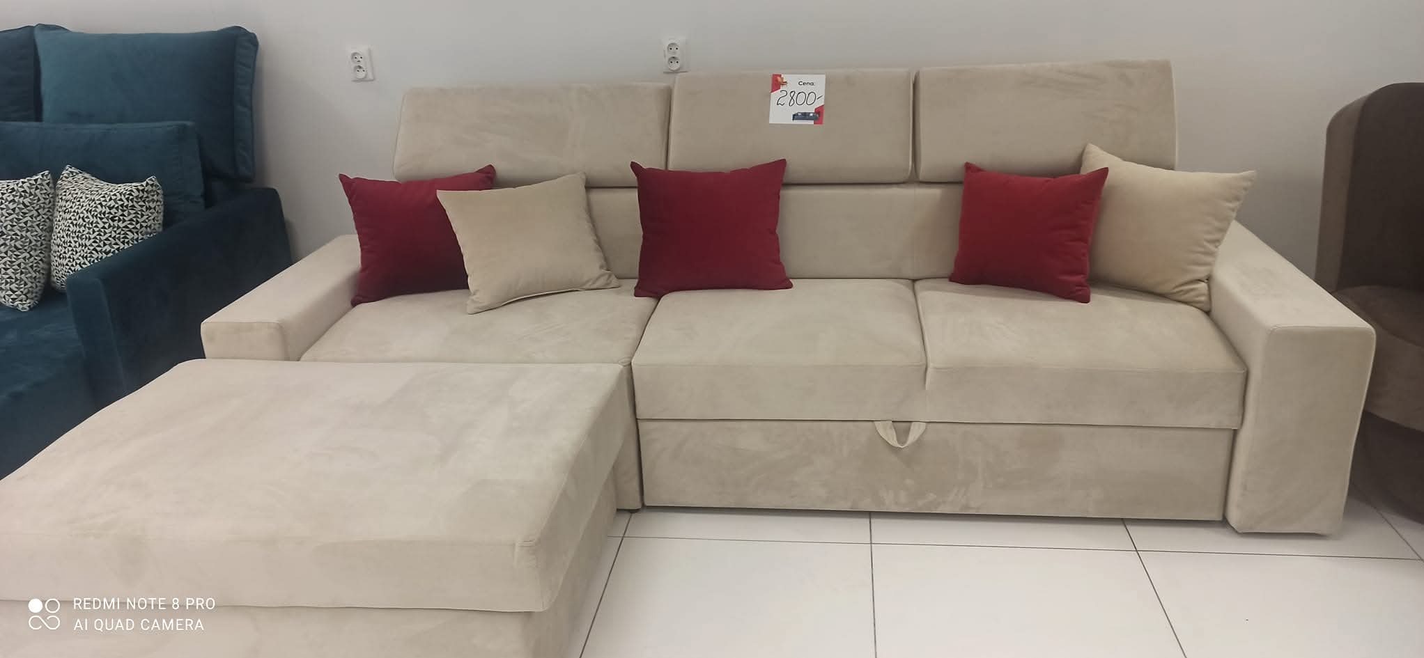 Beżowa sofa narożna z pufą i czerwonymi poduszkami, widoczna cena 2800 na kartce. Obok stoi ciemny fotel. Nowoczesny design, jasne tło.