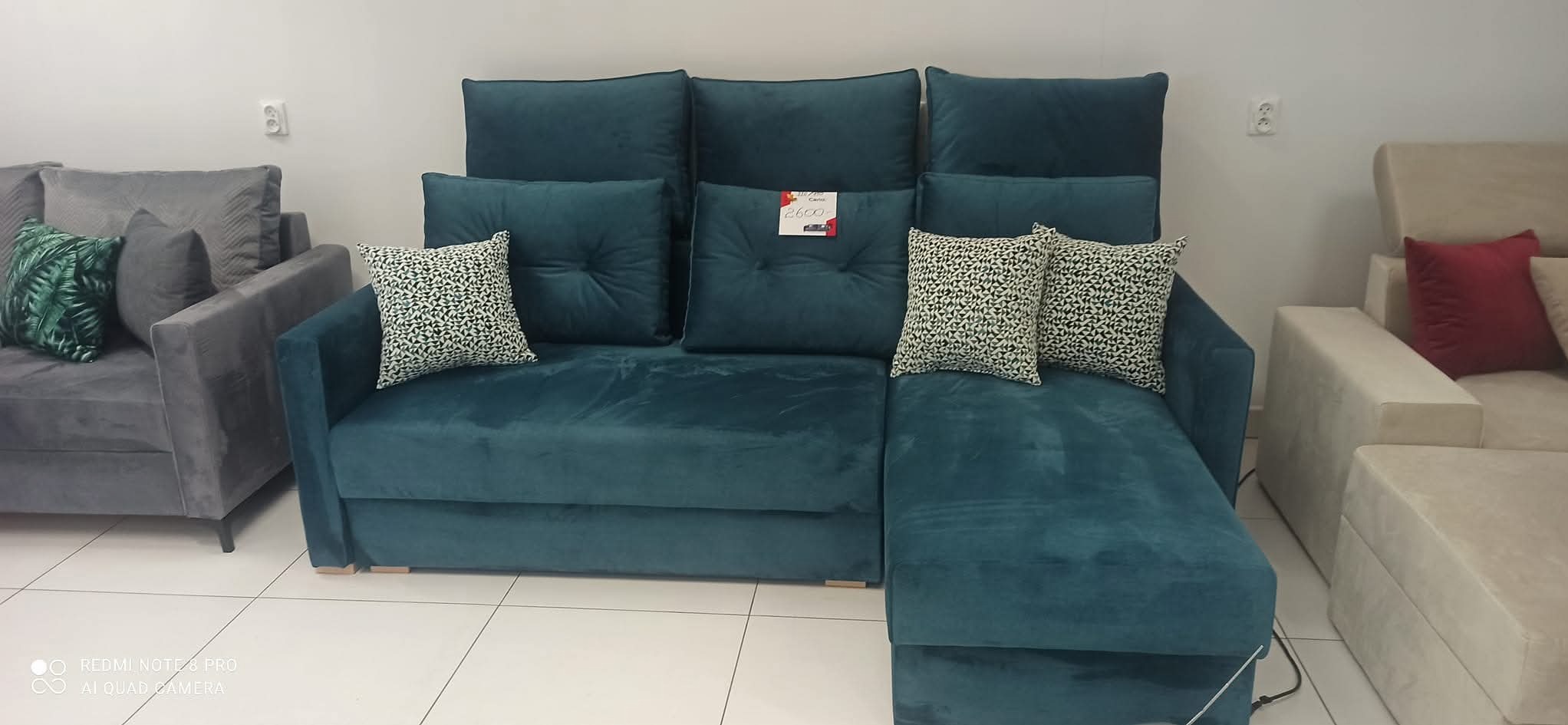 Turkusowa sofa z poduszkami w geometryczne wzory, widoczna w salonie. Na środkowej poduszce widoczna metka z ceną. Obok inne sofy w odcieniach szarości i beżu.