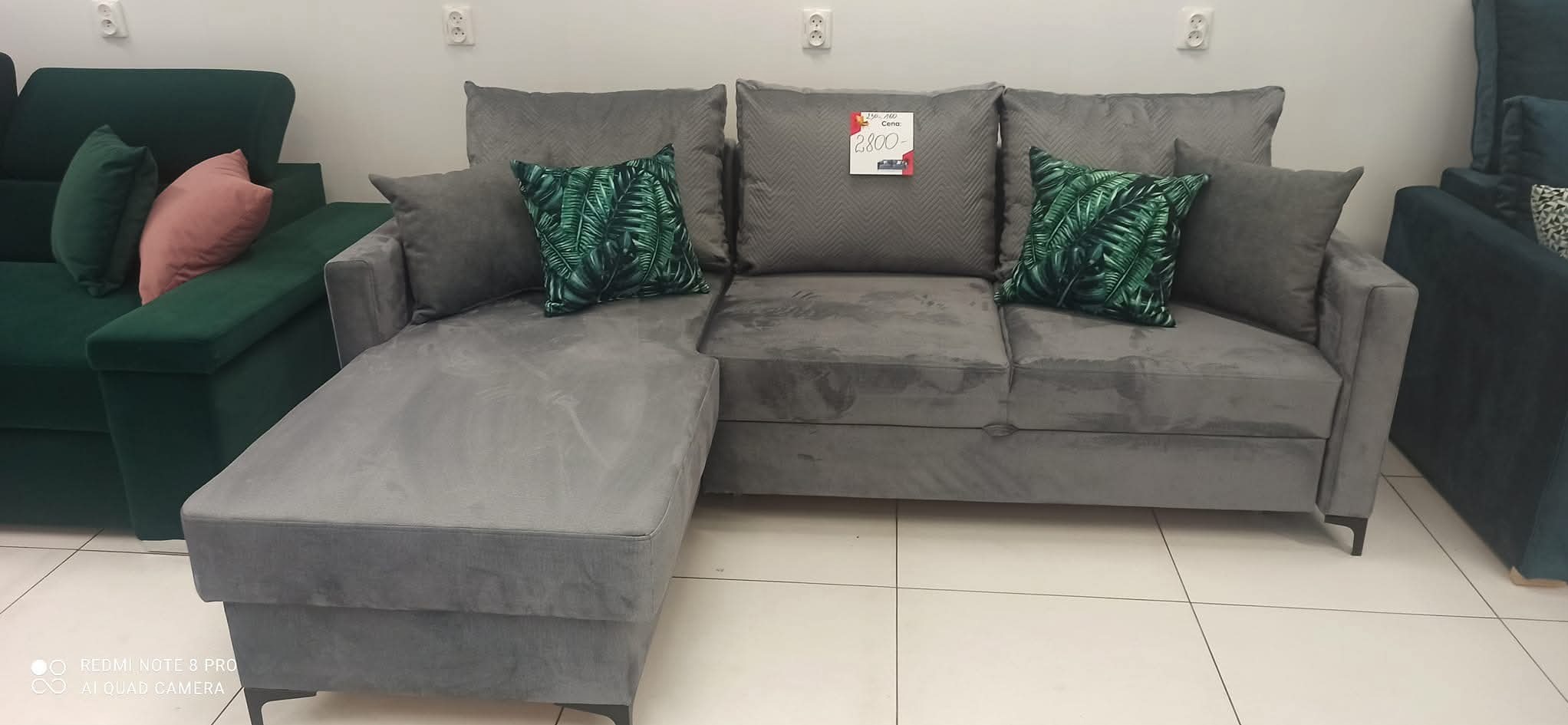 Szara sofa narożna z poduszkami w liście i ceną na jednej z nich, ustawiona w salonie meblowym, obok widoczny fragment zielonej sofy.