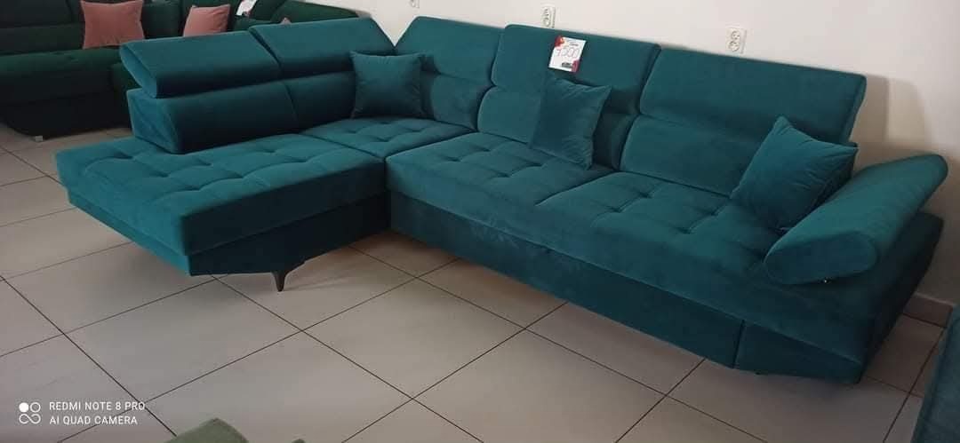 Turkusowy narożnik tapicerowany z poduszkami, widoczny w salonie meblowym. Narożnik z funkcją spania, pikowany, z zagłówkami. Na oparciu widoczna metka z ceną.