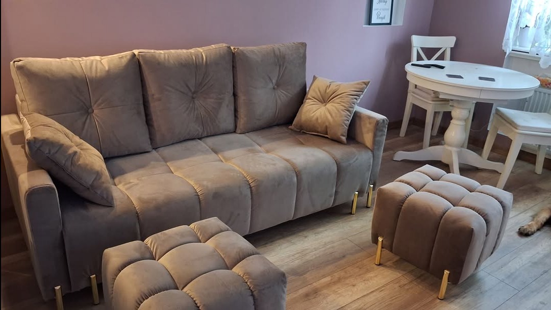 Beżowa sofa z poduszkami i dwoma pufami na złotych nóżkach w pokoju z białym stołem i krzesłami. Sofa w stylu Chesterfield z pikowaniem.