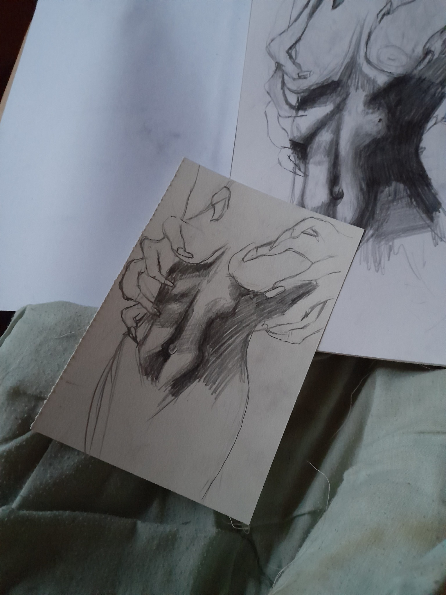 Szkic ołówkiem przedstawiający tors kobiety na kartce, obok drugi, większy szkic tego samego motywu, leżące na zielonej tkaninie.